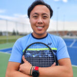 Coach Dominic holding a tennis racket on a tennis court on a sunny day.