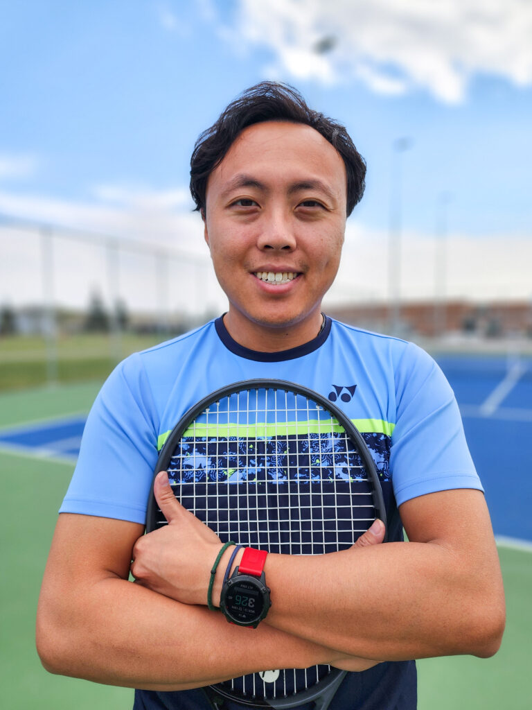 Coach Dominic holding a tennis racket on a tennis court on a sunny day.