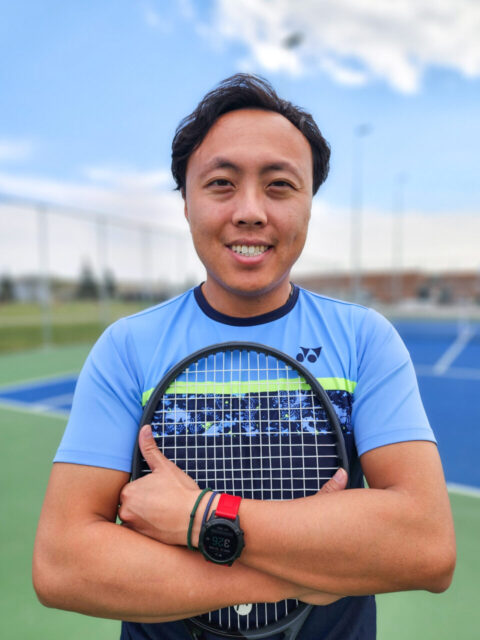 Coach Dominic holding a tennis racket on a tennis court on a sunny day.